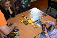 Dorośli i dzieci grający w gry planszowe przy bibliotecznych stolikach.