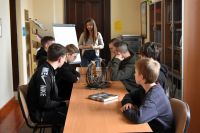 Grupa nastoletnich chłopców siedzi przy stolikach w bibliotece, słuchając stojącej przed nimi kobiety. W tle widać regały z...