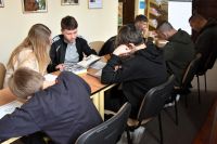 Grupa uczniów siedzi przy stolikach w bibliotece, skupiona na pracy, rozwiązują zadania na kartkach. W tle widać regały z...