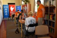 Grupa osób siedzi przy stolikach i gra w grę planszową. W tle regały z książkami i banery.