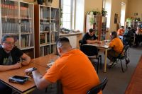 Grupa osób siedzi przy stolikach i gra w grę planszową. W tle regały z książkami i banery.