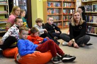 Grupa dzieci oraz dorosłych siedzi na pufach w bibliotece. W tle regały z książkami.
