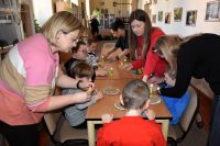 Grupa chłopców siedzi przy stolikach, stojące obok kobiety pomagają im w wykonaniu pracy plastycznej.