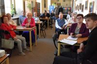 Grupa osób w różnym wieku siedzi przy stolikach w sali bibliotecznej