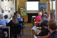 Uczestnicy spotkania siedzą przy stolikach w sali bibliotecznej i słuchają prezentacji. Na przodzie stoi kobieta prowadząca.