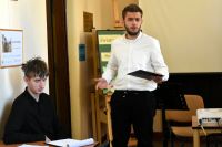 Dwóch młodych chłopców, jeden stoi i recytuje, drugi siedzi przy stoliku