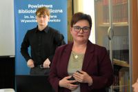 Kobieta stoi w sali bibliotecznej i przemawia, za nią w tle stoli młody chłopak