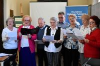 Grupa seniorów stoi w sali bibliotecznej i śpiewa