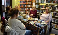 Kilka pań siedzi przy stoliku w sali bibliotecznej. Na stole leżą książki, talerz z ciastkami, filiżanki.