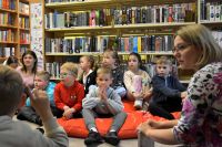 Grupa dzieci siedzi na pufach w bibliotece i słucha kobiety prowadzącej zajęcia. W tle widać regały pełne książek