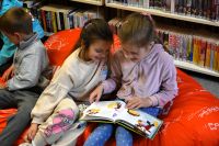 Dwie dziewczynki siedzą na pufie w bibliotece, oglądają wspólnie książkę.