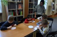Grupa dzieci siedzi przy stolikach w bibliotece, kolorują papierowe kółka.