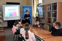 Grupa dzieci siedzi przy stolikach w bibliotece. W tle kobieta prowadząca zajęcia oraz ekran z prezentacją.