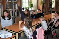 Grupa dzieci siedzi przy stolikach w bibliotece. Dziewczynka podnosi rękę.