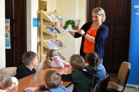 Grupa dzieci siedzi przy stolikach w bibliotece. Słuchają kobiety, która opowiada i pokazuje książkę.
