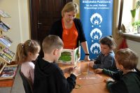 Grupa dzieci siedzi w bibliotece przy stolikach, nawlekają obrazki na sznurek. Przy stoliku stoi kobieta z książką.