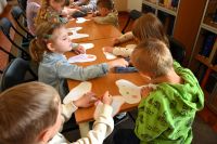 Grupa dzieci siedzi przy stolikach w bibliotece i kolorują papierowe motyle.