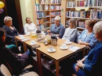Grupa kobiet, siedząca przy stolikach. Na stolikach filiżanki, ciastka, książki