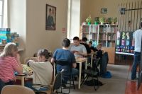 Uczestnicy spotkania Planszówki w bibliotece, dzieci i osoby dorosłe siedzą przy stolikach i grają w gry planszowe