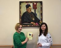 Dwie bibliotekarki na tle ściany z obrazem, przedstawiającym patrona biblioteki .