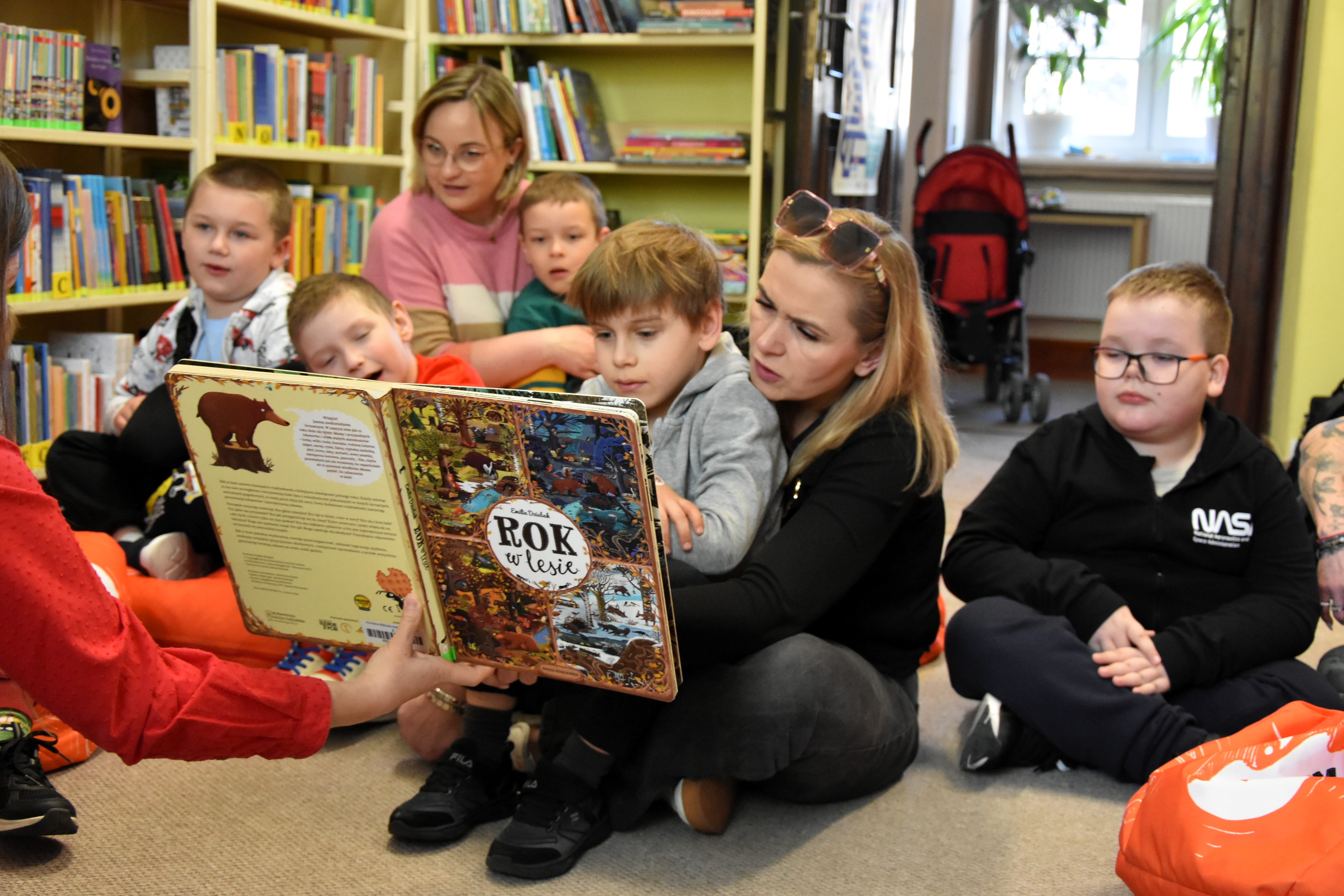 Grupa dzieci siedzi na pufach w bibliotece i ogląda książkę ,,Rok w lesie".