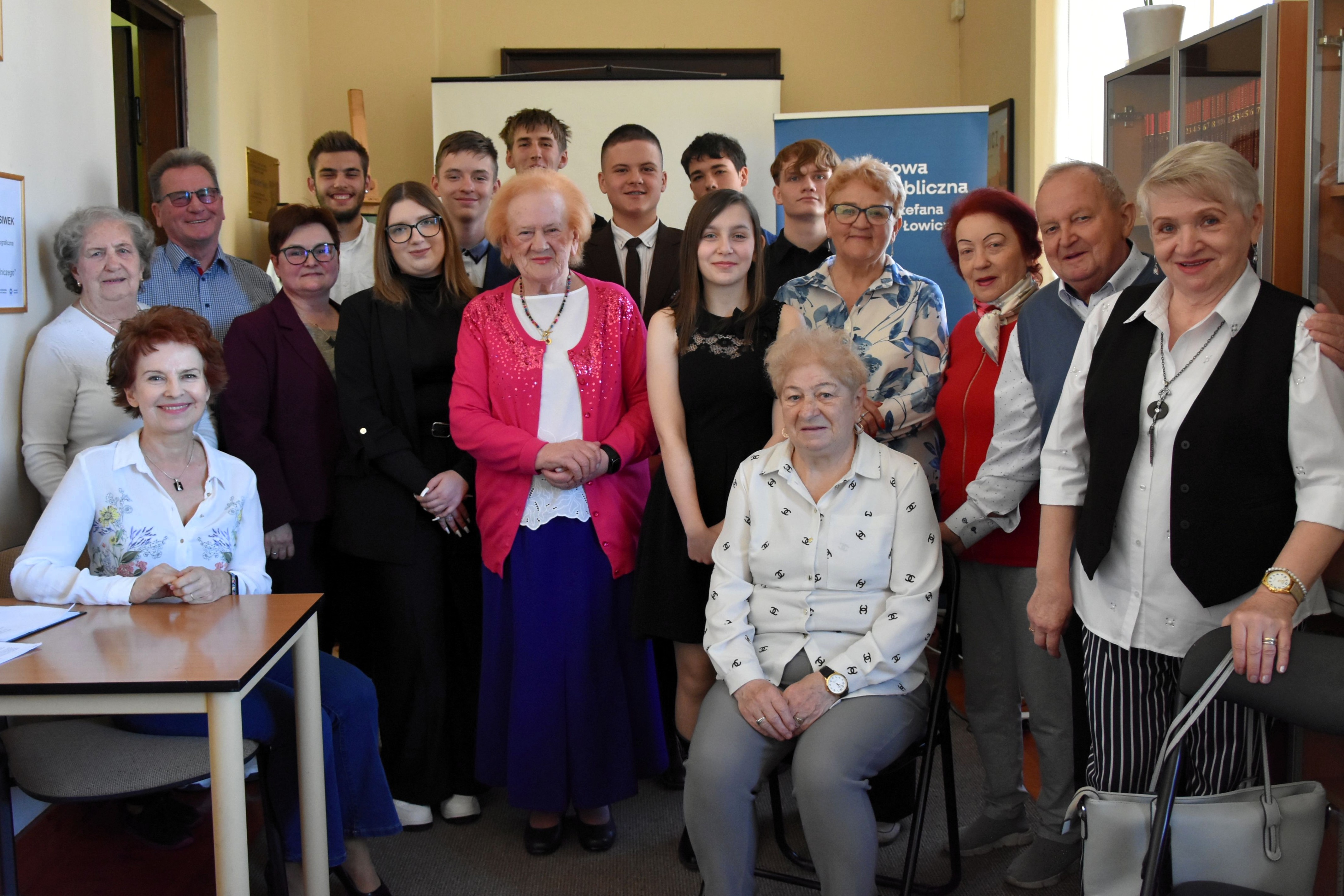 Grupa osób w różnym wieku pozuje do wspólnego zdjęcia w sali bibliotecznej