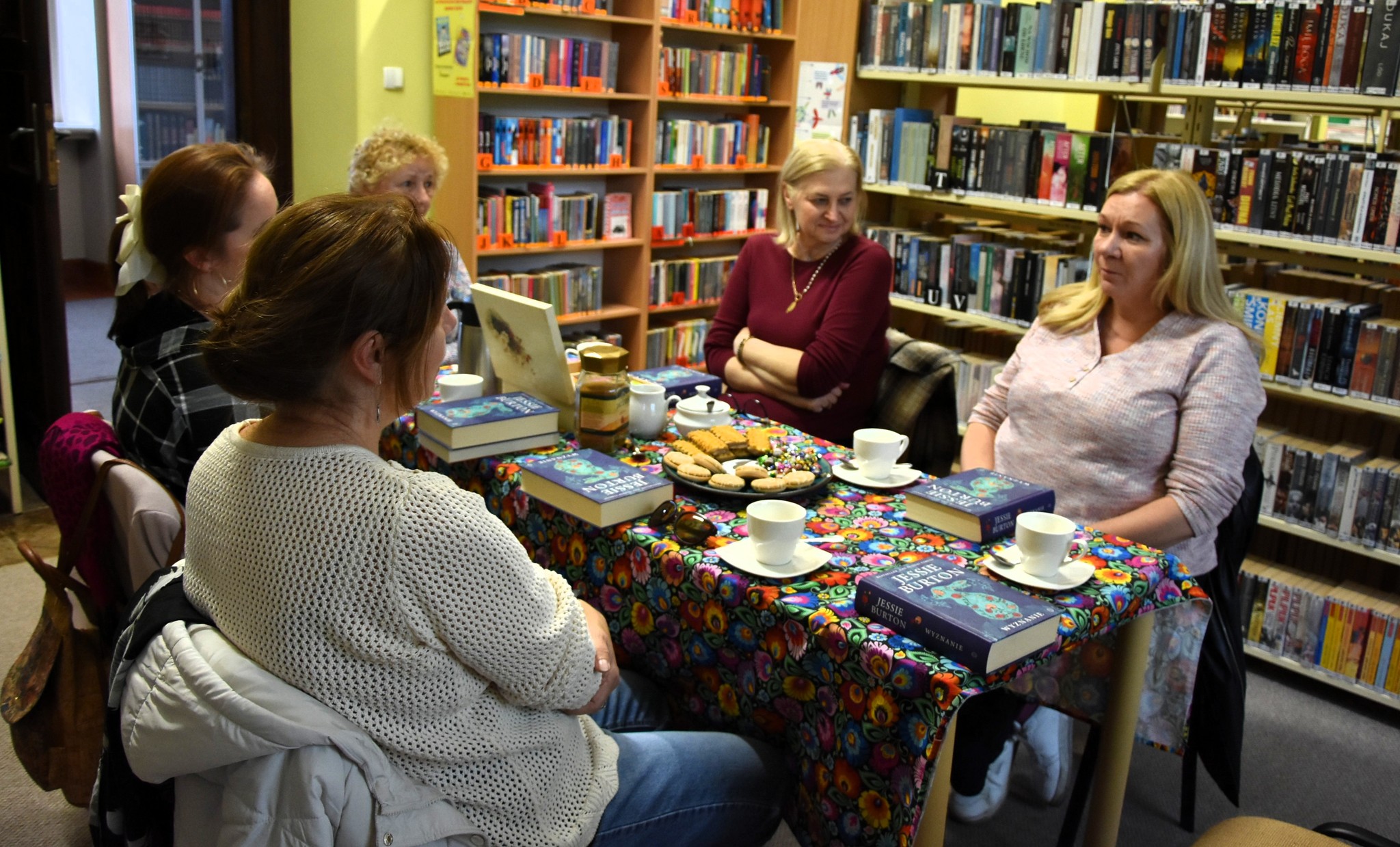 Kilka pań siedzi przy stoliku w sali bibliotecznej. Na stole leżą książki, stoi talerz z ciastkami oraz filiżanki.